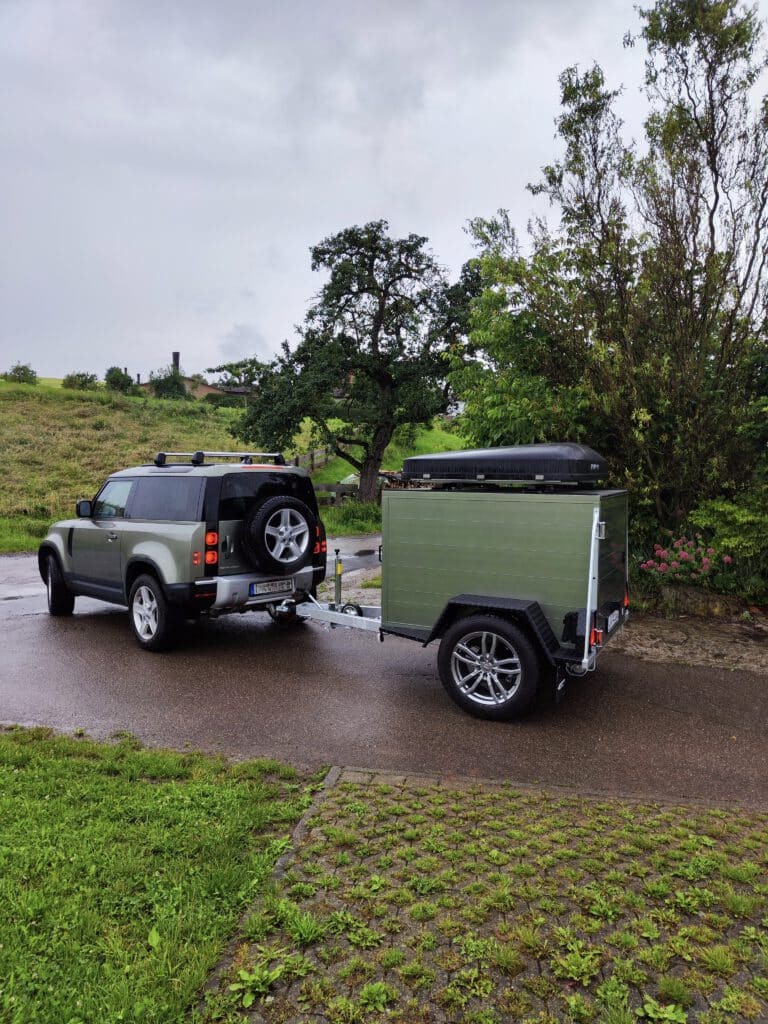 Der Offroadanhänger Corse setzt neue Maßstäbe für Abenteurer, die maximale Freiheit und Funktionalität suchen. Als hochwertiger Zeltanhänger wurde er speziell für anspruchsvolle Touren entwickelt und überzeugt durch robuste Bauweise, intelligente Ausstattung und modernes Design. Die seitliche Tür sorgt für einen besonders komfortablen Zugang zum Innenraum und ermöglicht eine flexible Nutzung des Stausystems – ideal für Camping, Expeditionen oder Wochenendtrips. Dank der integrierten Kugelaufnahme für Fahrradträger lässt sich der Offroadanhänger Corse problemlos um zusätzliche Transportkapazität erweitern. Perfekt für alle, die ihre Bikes auch abseits befestigter Wege einsetzen möchten. Die stabile Reling R.O.Rack eignet sich optimal zur Montage eines Dachzelts und verwandelt den Offroadanhänger in eine vollwertige Outdoor-Base für mehrtägige Abenteuer. Optisch besticht der Anhänger mit einer exklusiven Lackierung in Land Rover Pangea Grün – ein echter Hingucker, der Robustheit und Stil vereint. Für maximale Unabhängigkeit verfügt der Zeltanhänger über eine vollständige 230-Volt-Elektroinstallation, die sämtliche Camping- und Ladegeräte zuverlässig versorgt. Ergänzt wird das Setup durch moderne LED-Fahrzeugbeleuchtung, die für beste Sichtbarkeit und maximale Energieeffizienz sorgt – selbst unter härtesten Offroad-Bedingungen.
