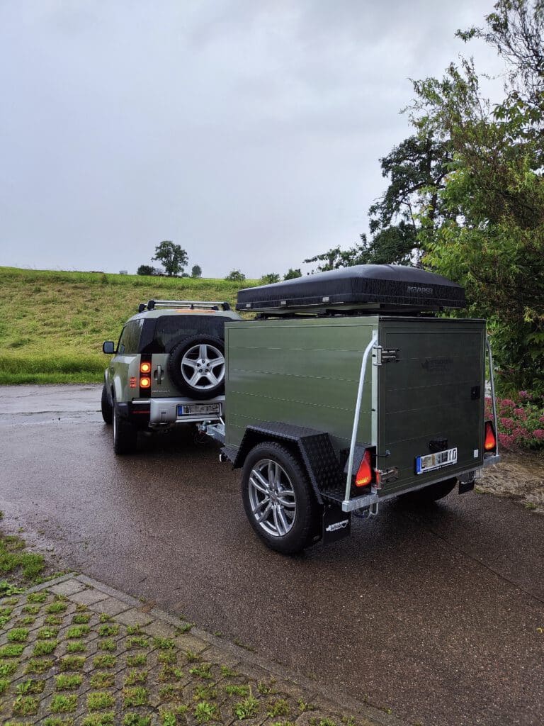Der Offroadanhänger Corse setzt neue Maßstäbe für Abenteurer, die maximale Freiheit und Funktionalität suchen. Als hochwertiger Zeltanhänger wurde er speziell für anspruchsvolle Touren entwickelt und überzeugt durch robuste Bauweise, intelligente Ausstattung und modernes Design. Die seitliche Tür sorgt für einen besonders komfortablen Zugang zum Innenraum und ermöglicht eine flexible Nutzung des Stausystems – ideal für Camping, Expeditionen oder Wochenendtrips. Dank der integrierten Kugelaufnahme für Fahrradträger lässt sich der Offroadanhänger Corse problemlos um zusätzliche Transportkapazität erweitern. Perfekt für alle, die ihre Bikes auch abseits befestigter Wege einsetzen möchten. Die stabile Reling R.O.Rack eignet sich optimal zur Montage eines Dachzelts und verwandelt den Offroadanhänger in eine vollwertige Outdoor-Base für mehrtägige Abenteuer. Optisch besticht der Anhänger mit einer exklusiven Lackierung in Land Rover Pangea Grün – ein echter Hingucker, der Robustheit und Stil vereint. Für maximale Unabhängigkeit verfügt der Zeltanhänger über eine vollständige 230-Volt-Elektroinstallation, die sämtliche Camping- und Ladegeräte zuverlässig versorgt. Ergänzt wird das Setup durch moderne LED-Fahrzeugbeleuchtung, die für beste Sichtbarkeit und maximale Energieeffizienz sorgt – selbst unter härtesten Offroad-Bedingungen.